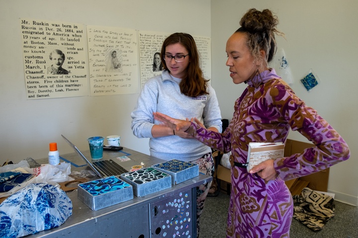 Two people stand next to a table with art pieces displayed. One gestures to the work and speaks while the other listens.