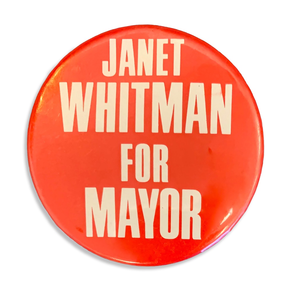 A large red pin against a white background has the words “JANET WHITMAN FOR MAYOR” printed in an off-white color.