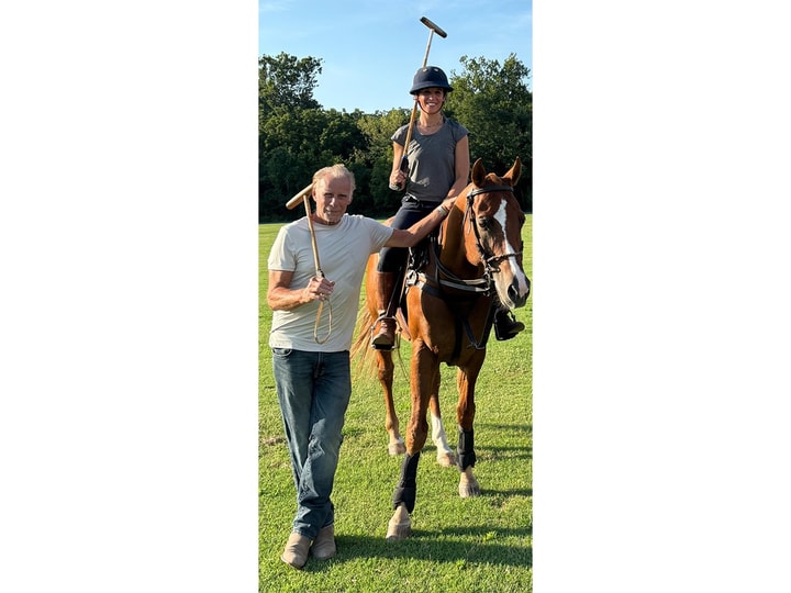 A man holding a polo mallet over one shoulder stands next to a horse with a woman sitting atop in polo gear.