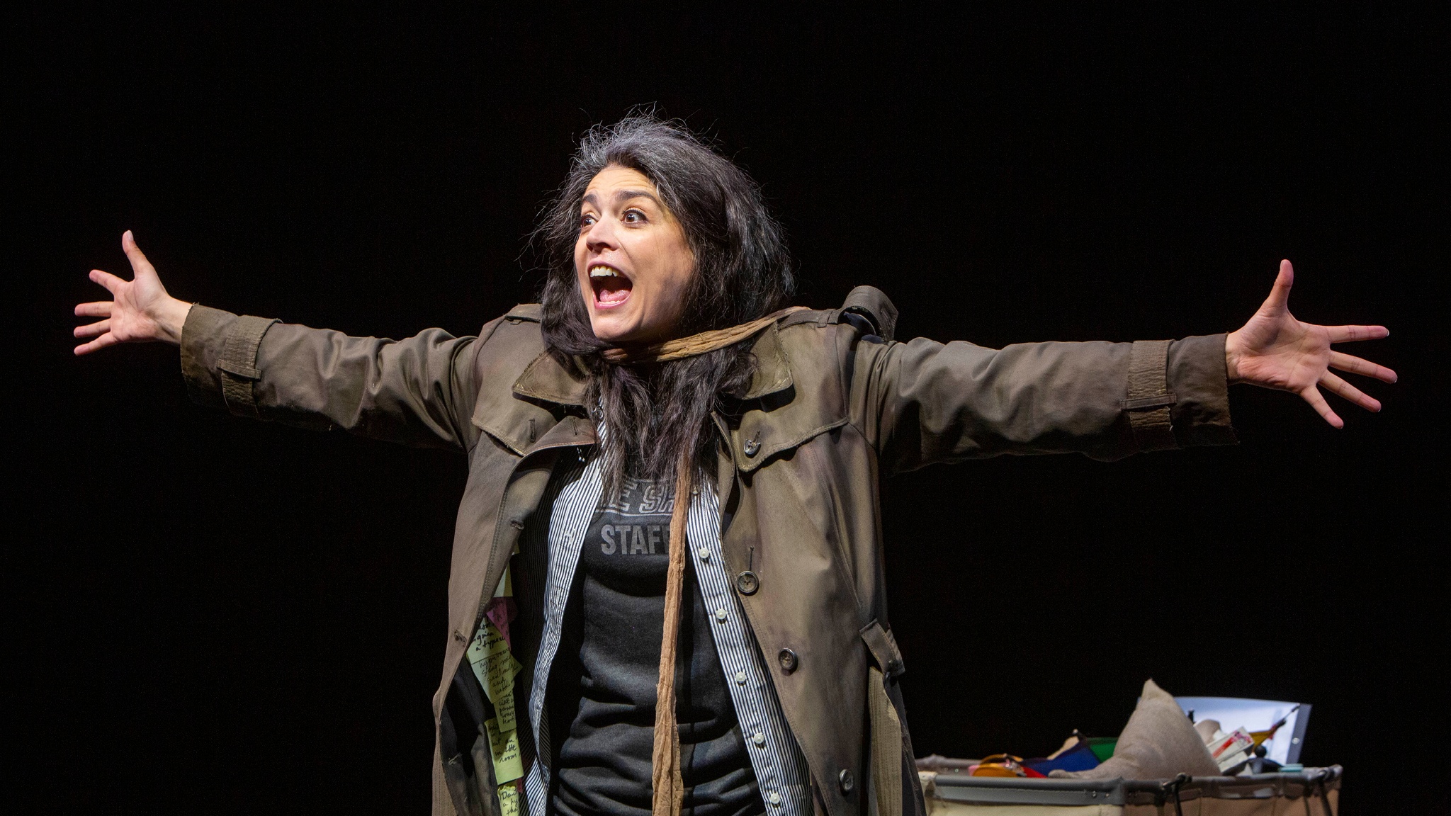 The performer Cecily Strong on a black box theater stage wearing a wig with graying hair, a brown trench coat, and holding her arms outstretched