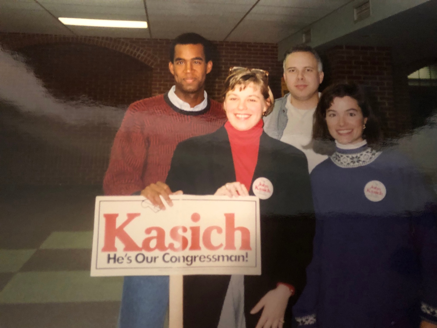 Four adults standing together inside a brick interior are smiling at the camera. The two women in front are wearing pins and holding a picket sign that says, “Kasich He’s Our Congressman!”