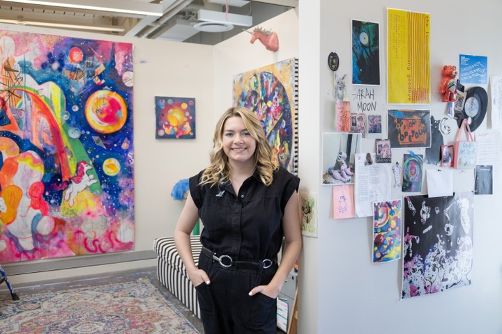 A woman with blonde hair wearing black leaning against the corner of two walls covered in bright colorful art 