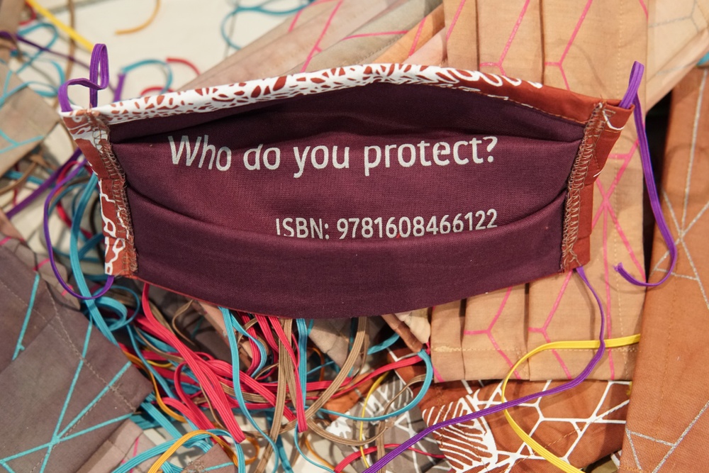 The back of a maroon, handmade mask with white text printed on it reading "Who do you protect? ISBN: 9781608466122."