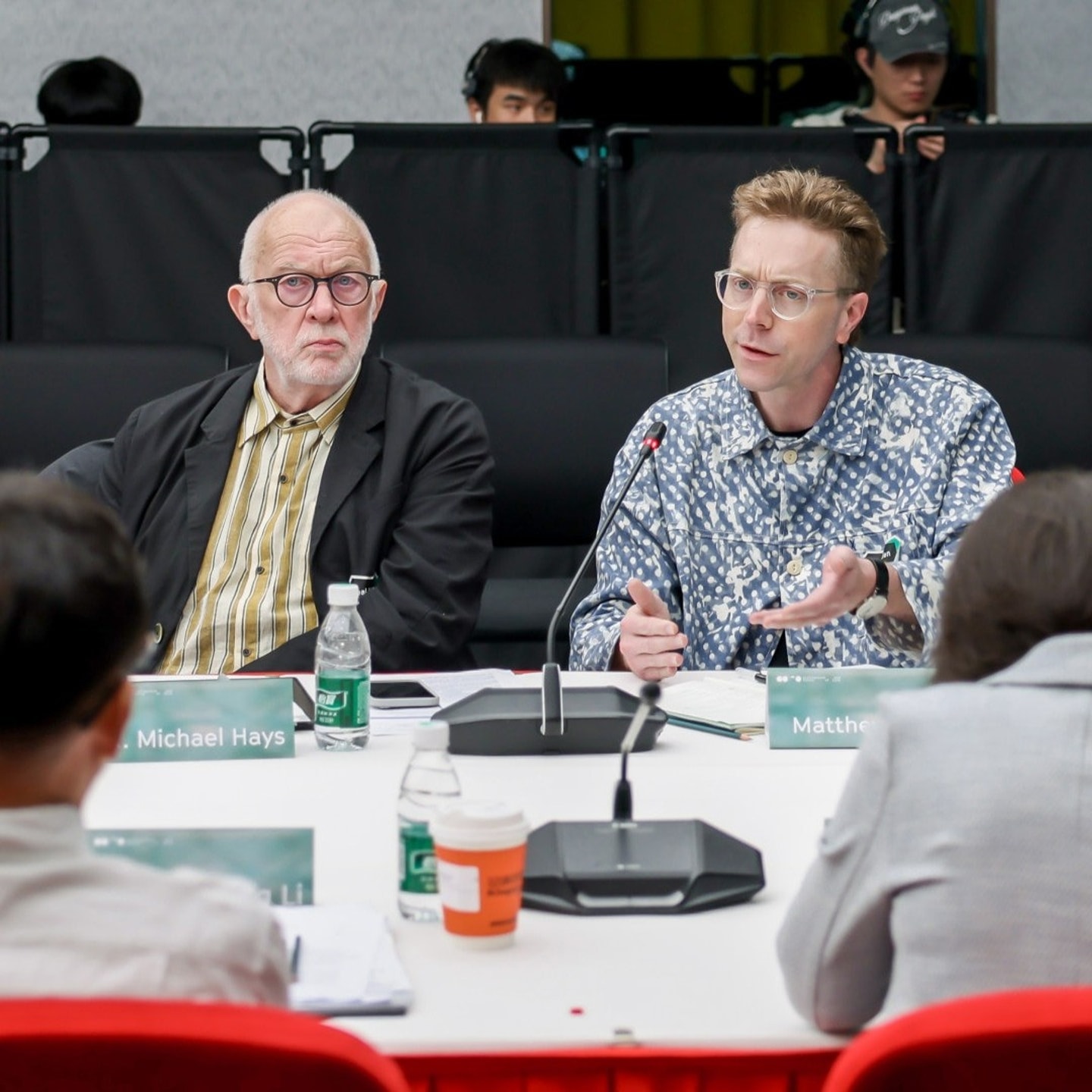 Two men seated at a conference table, one listening and one speaking, looking at two people on the other side of the table.