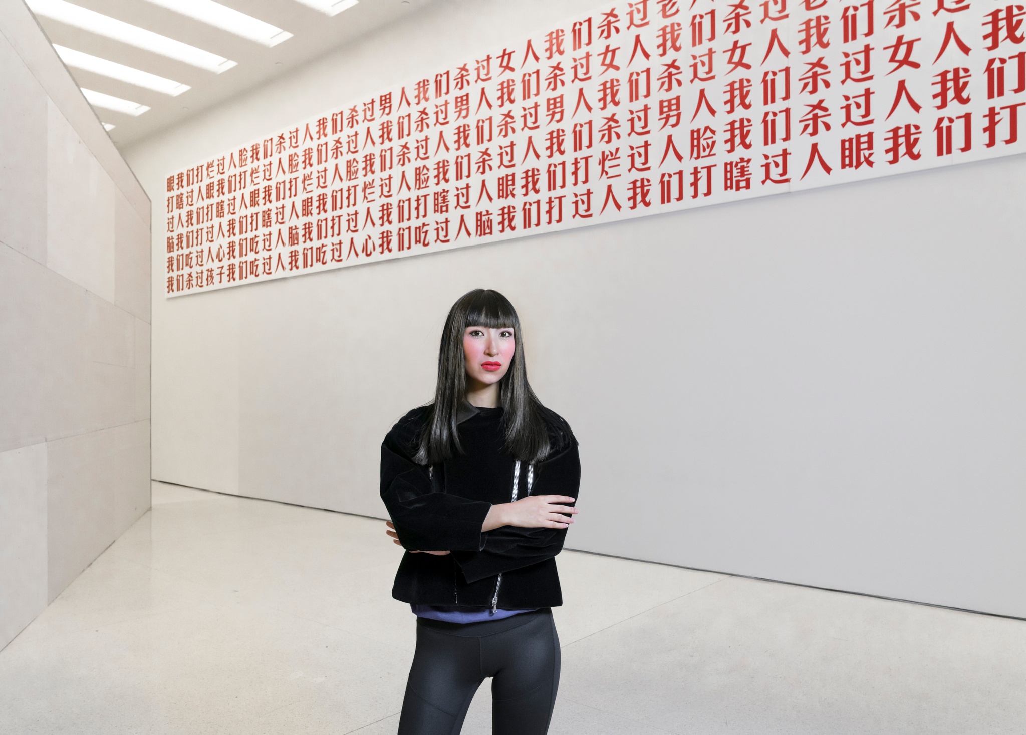 A photo of X Zhu-Nowell in a black jacket and with arms crossed, posing in front of a gallery installation with red Chinese characters on the wall. 