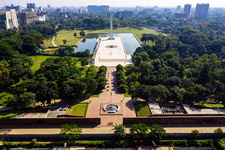 Aerial photo of an open plaza area with water on the sides, surrounded by dense trees; a city skyline is in the background.