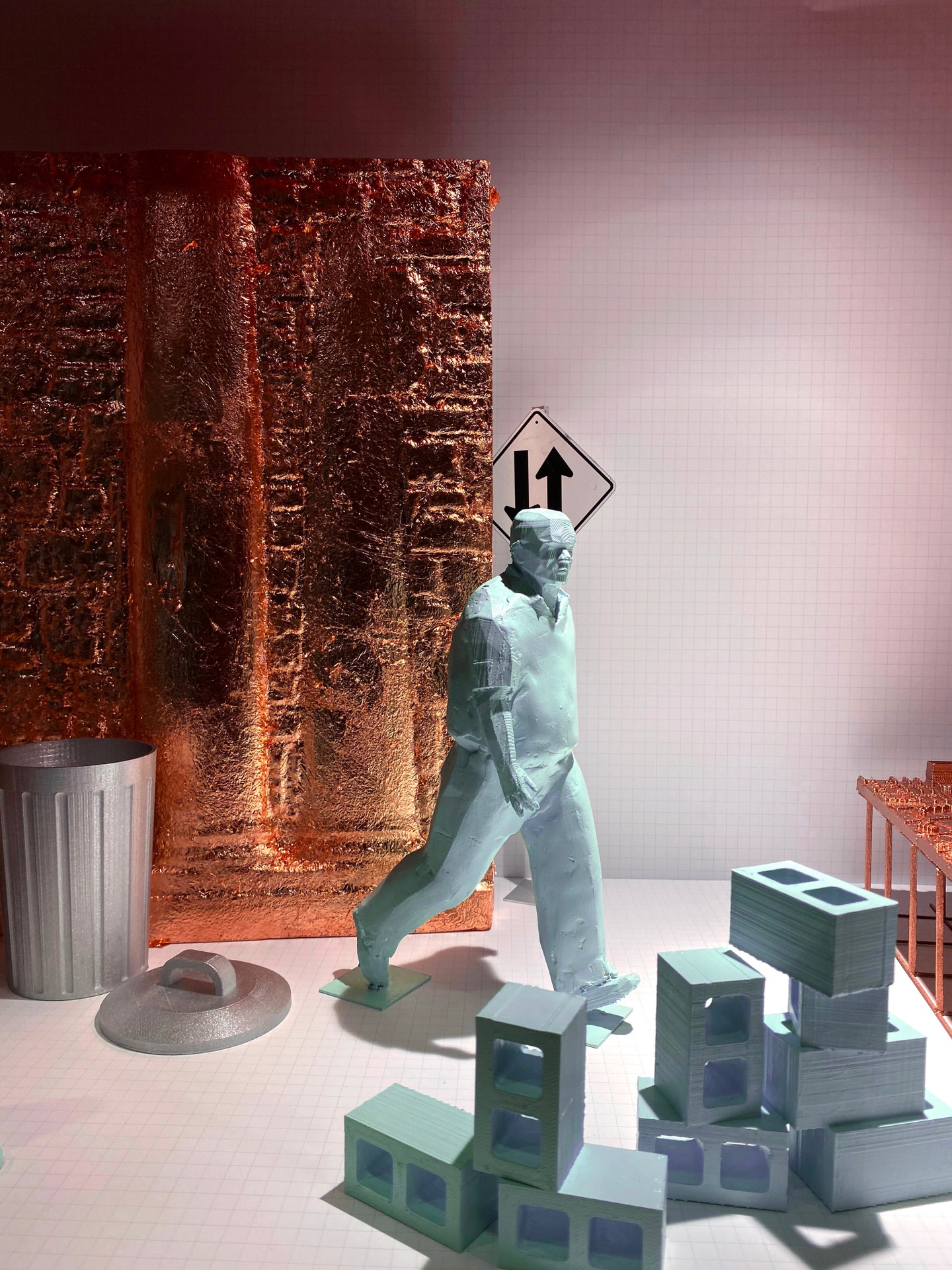 Detail of an architectural model featuring a sculpted figure walking in front of a pile of cinder blocks, with a trash can and part of a wall behind the figure.