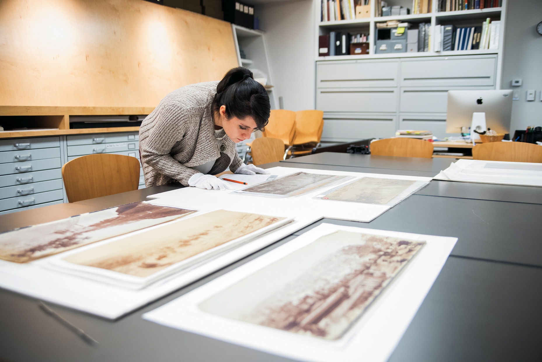 Ana-Joel Falcon-Wiebe, a light-skinned female, examines sepia-colored photographs depicting desert landscapes while wearing white gloves.