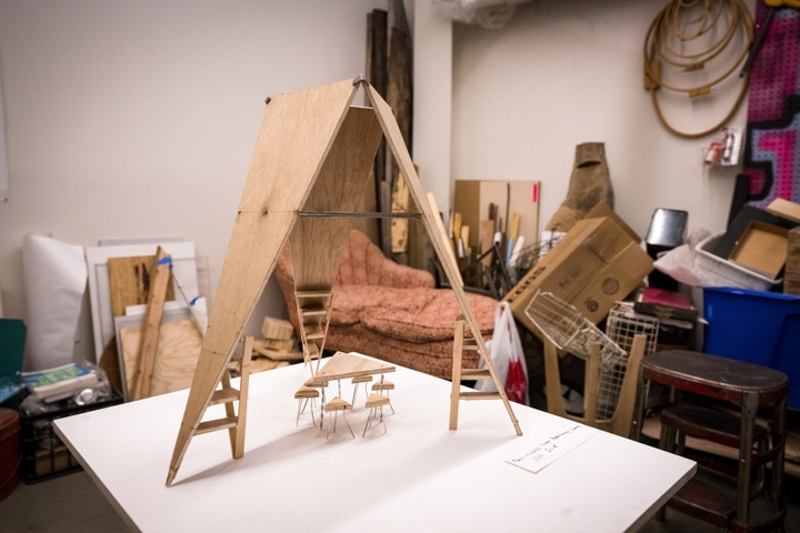 A small white-topped table stands in the middle of a cluttered studio space. On the table is a tiny wooden model of a triangular table and triangular stools sitting underneath a canopy constructed of three tall triangles of wood leaned end to end against each other, pointy tips on the table top.