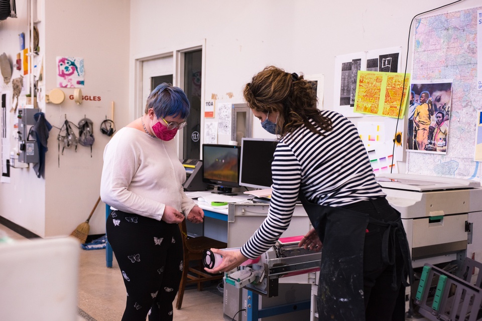 A person with short blue hair watches while a person in a black apron changes out a pink ink drum in the risograph machine. Colorful prints and safety goggles are hung on the wall behind them.