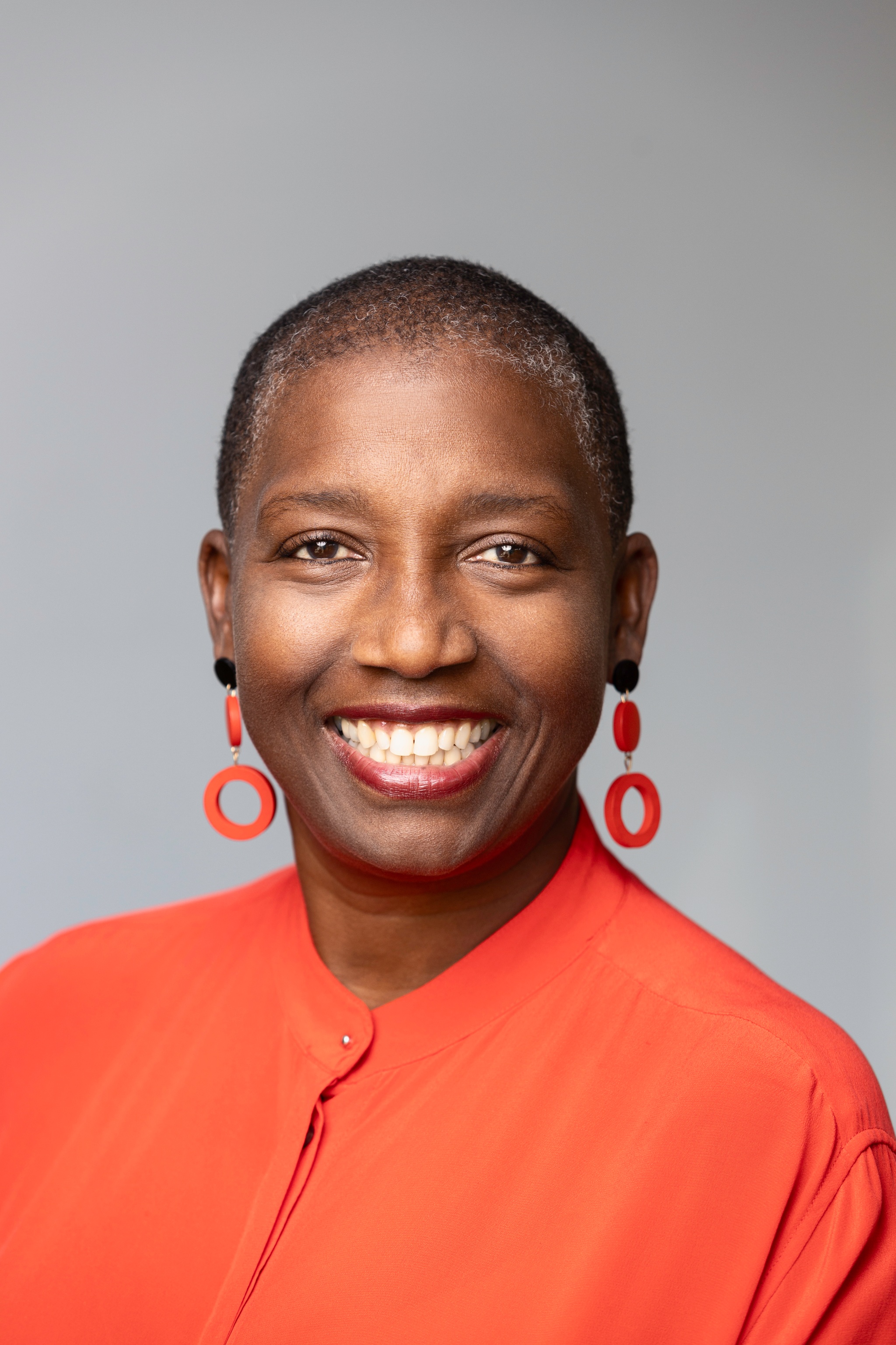 A portrait of Saundra Thomas, a Black woman with hair shaved to her head. She smiled broadly and looks directly at us. She wears dangling orange earrings that match her orange blouse. 