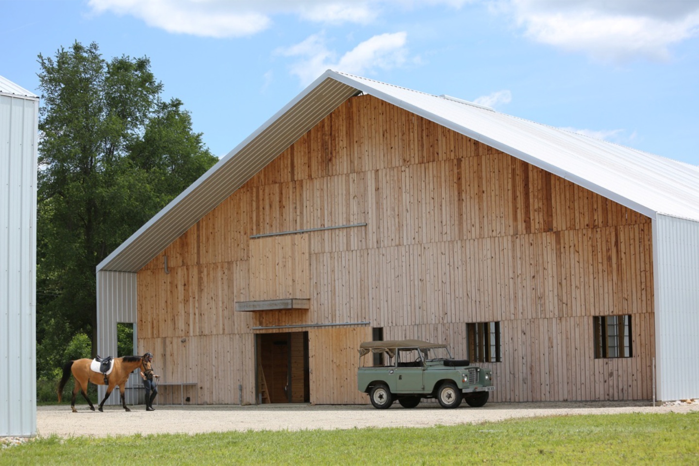 Front of a gabled barn with white roof and siding overhanging the entryway. The front wall is constructed of four rows of vertical wood slats and includes a sliding barn door and two small windows.