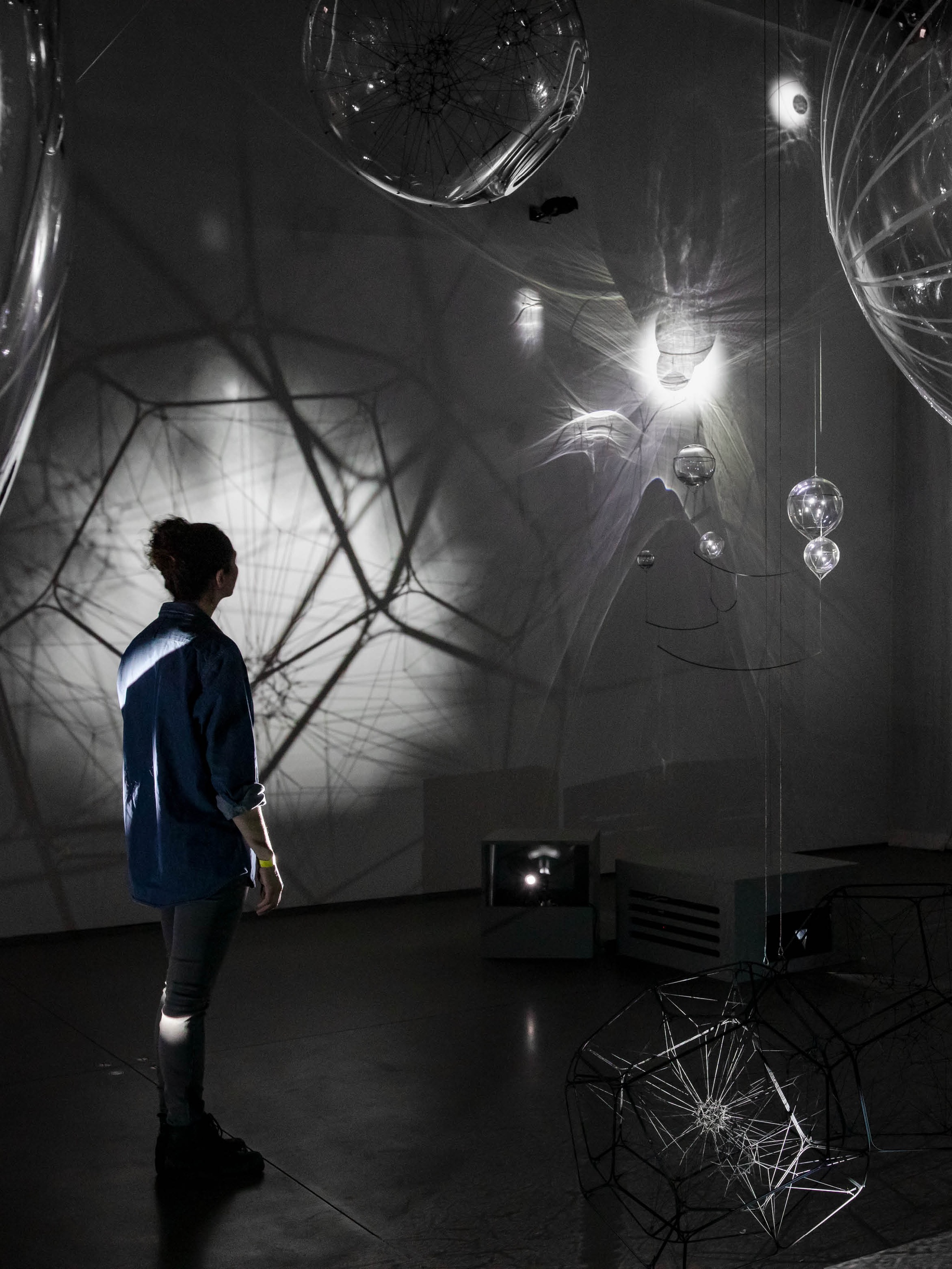 A person stands in shadowy light in an gallery with light refracted through hanging orbs and other sculptures
