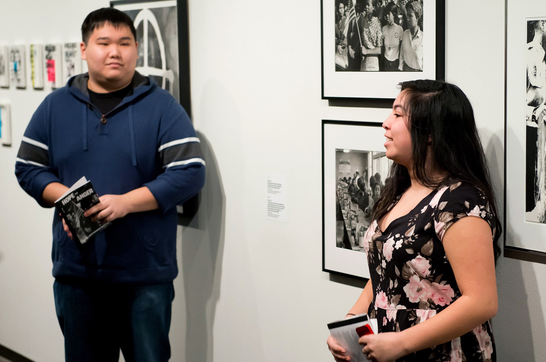 Two light-skinned, young people, one male and one female, stand in front of a wall with black and white photographs.