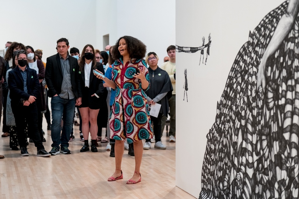 Person stands in front of a crowd in a gallery space and speaks. Behind them is the edge of a large-scale black and white drawing of a twisted fabric skirt.