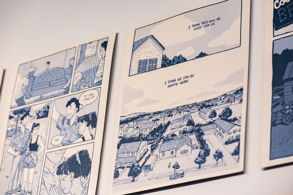 Close up on a spread of comics pages mounted on a wall. The drawings are in shades of dark blue and shows scenes of a family moving into a house. Narrative text reads "I think this will be good for us. I think we can be happy here."