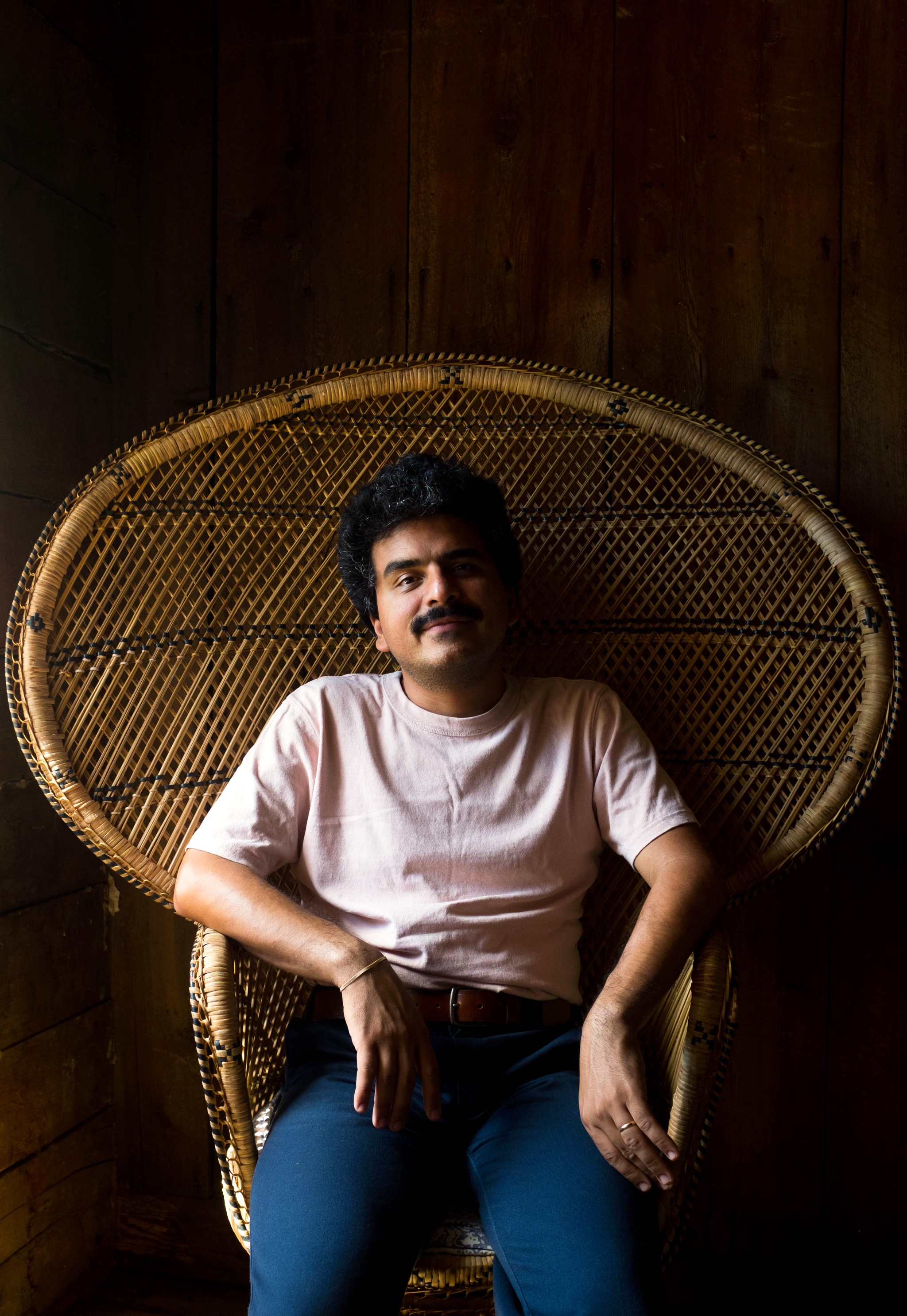 A photo of Roberto Carlos Lange sitting in light and shadow in a wide-backed wicker chair. 