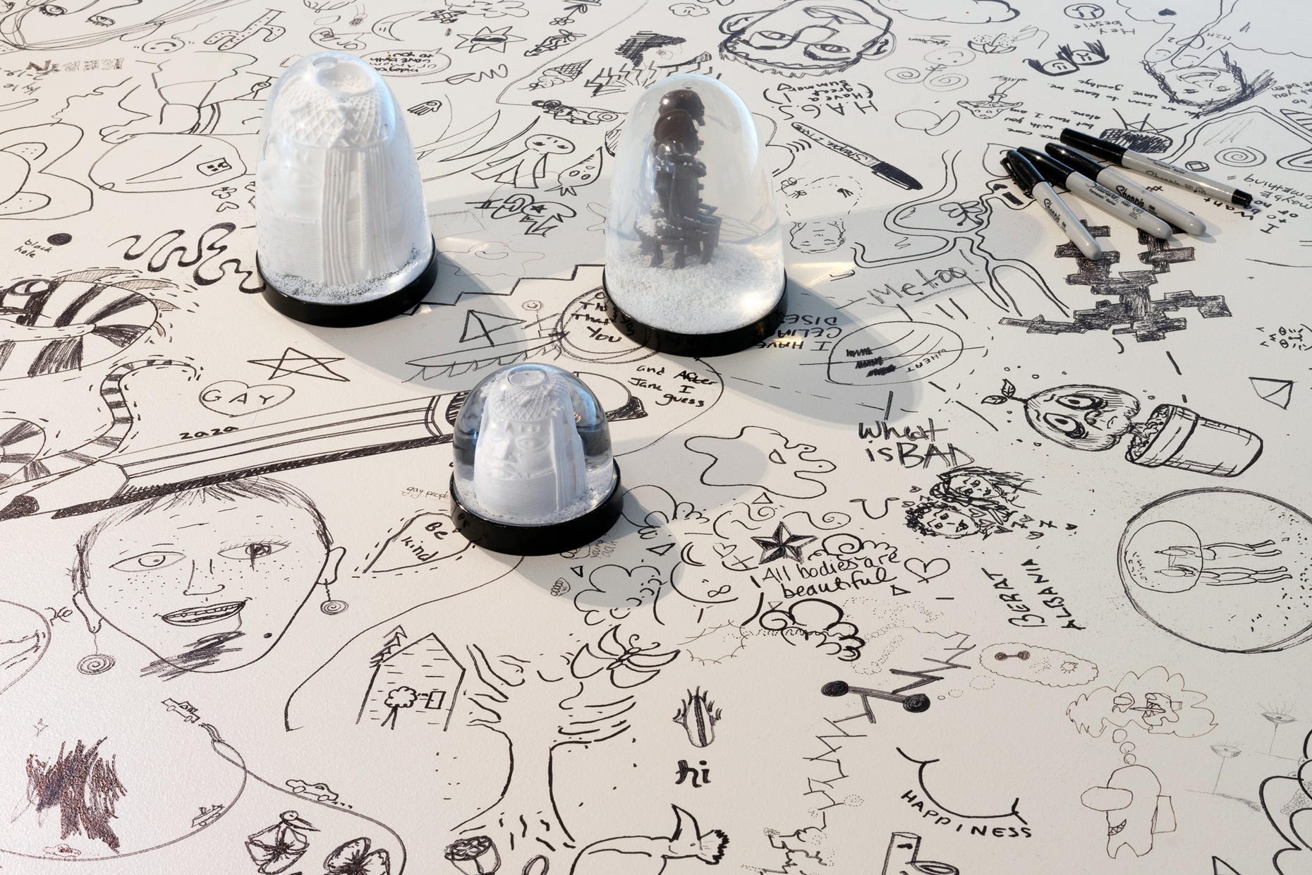 On a white surface are black drawings and doodles, along with various blurbs of handwritten text, atop which sits three black and white snow globes and four sharpies.