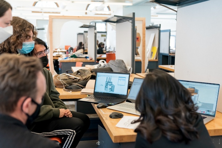 Group of people huddle around three laptops side by side on a desk, displaying digital renderings of machinery.