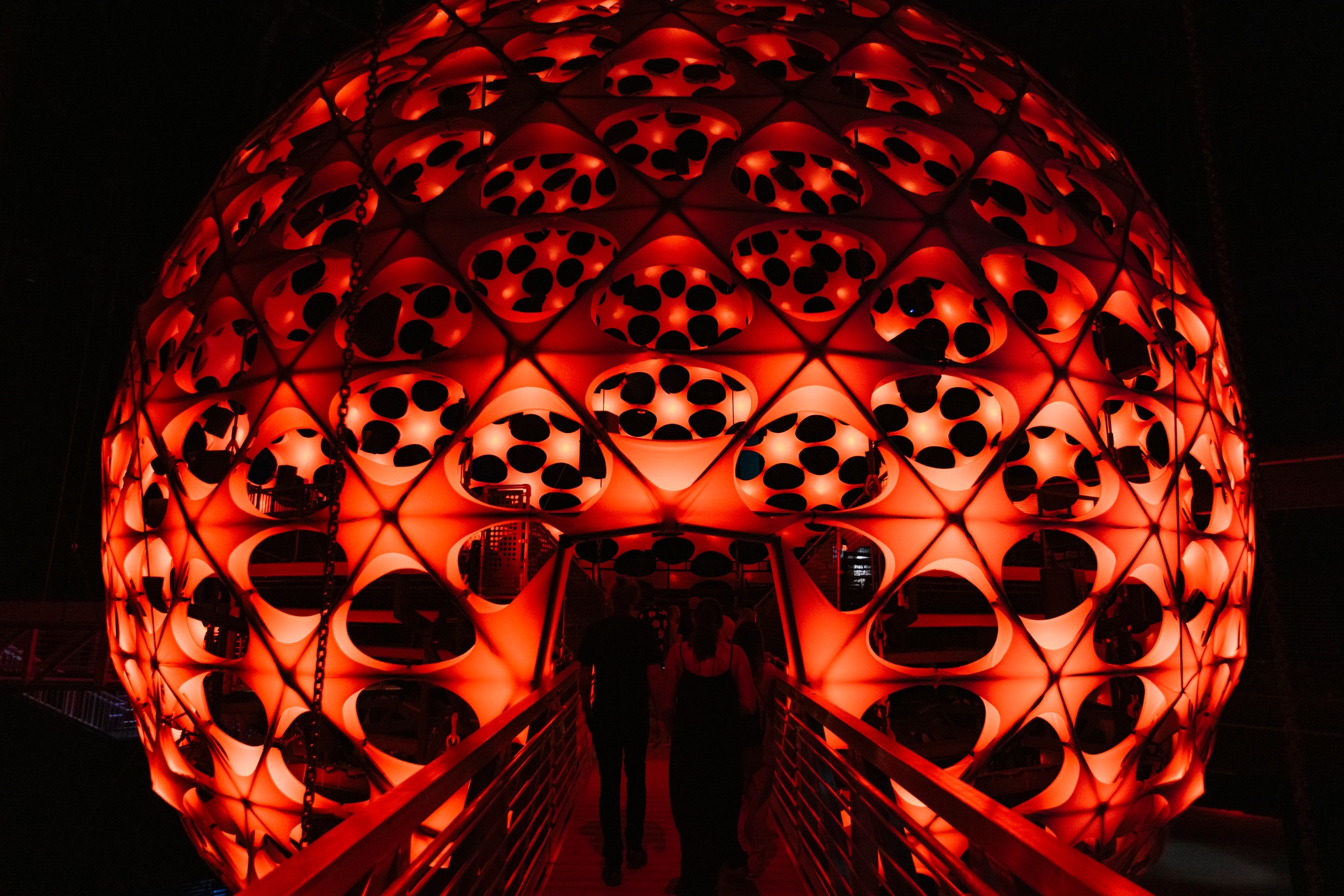 Two people seen from behind enter a large spherical concert hall. The surface of the sphere is dotted with light nodes that emanate a red-orange glow.