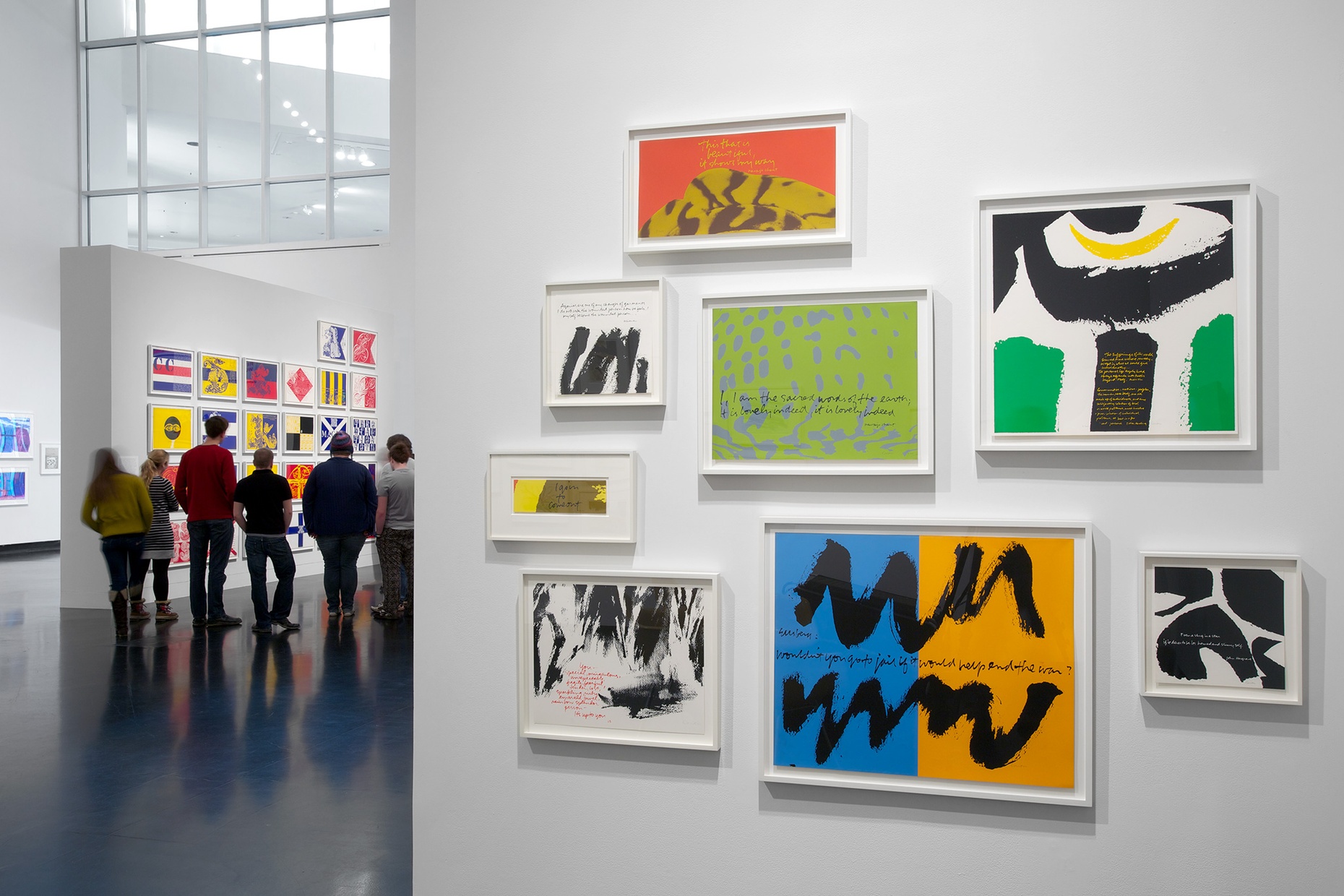Eight colorful screen prints hung on a white wall with a group of people visible in the background.