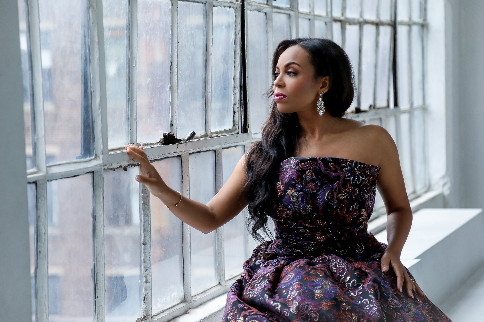 A Black woman dressed in a purple floral print shoulderless gown. She sits on a windowsill looking out with one hand resting on the windowpane. 
