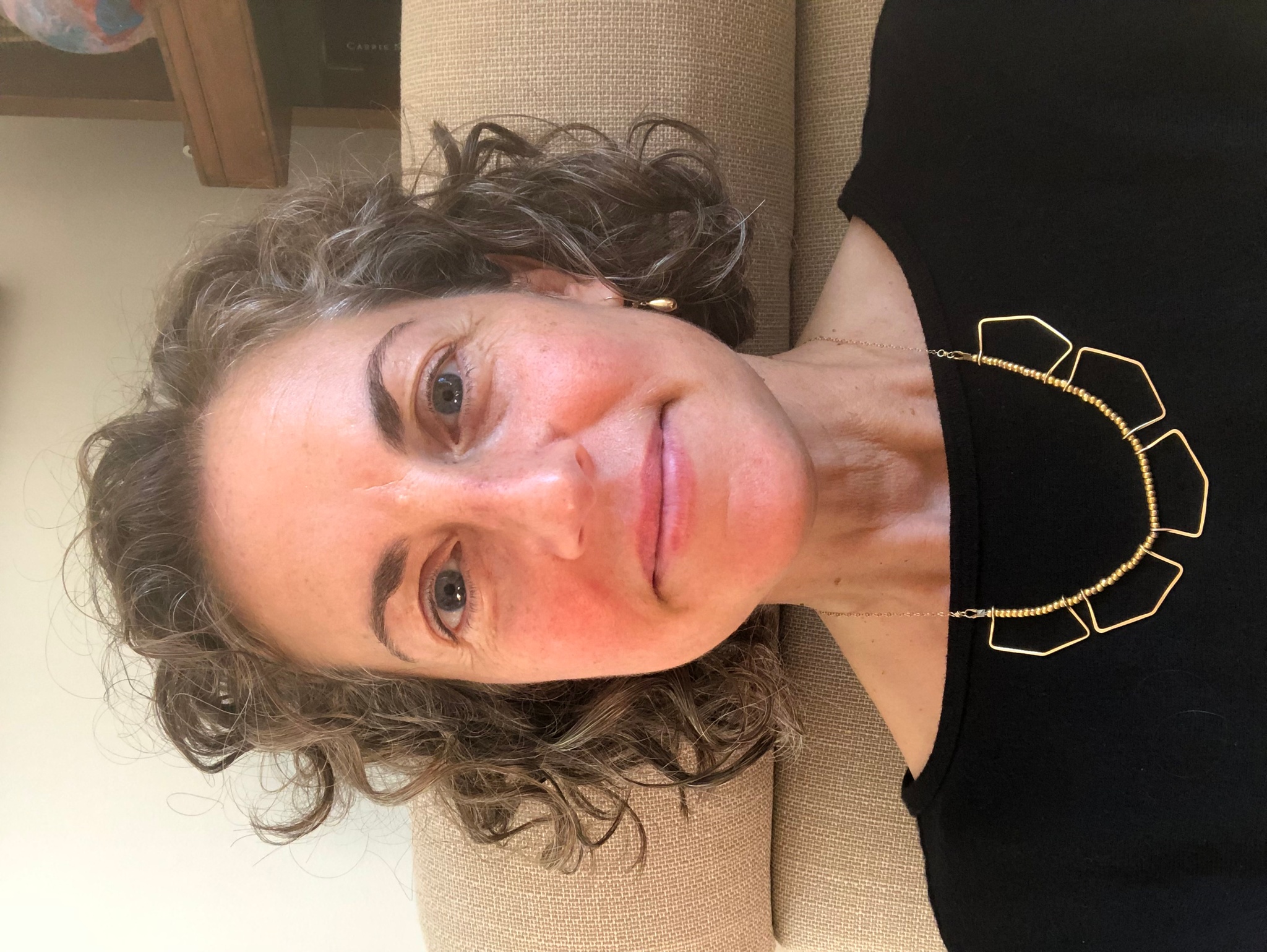 A portrait of Nadine Goellner, a white woman who smiles with mouth closed and eyebrows raised She has shortish curly brown and gray hair and wears a metal necklace with loops on top of a black boatneck shirt. 