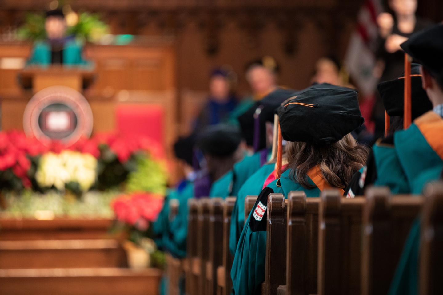 Rows of people in green graduation gowns with black mortarboards, seated in brown pews; there's a soft focus on the background, where someone in cap and gown stands at a podium.