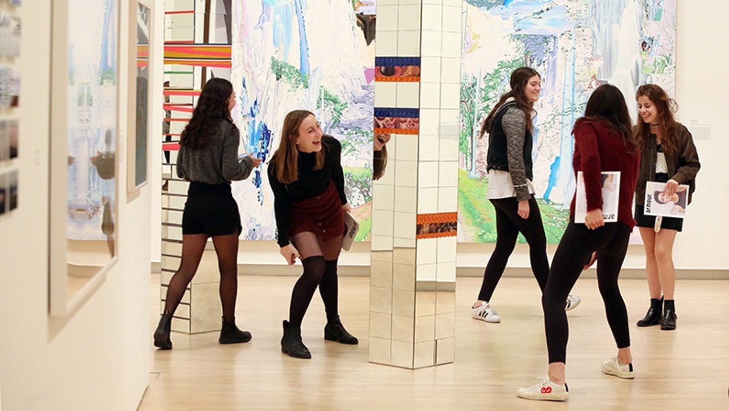 Five young female-presenting people laugh and play around artworks in a gallery