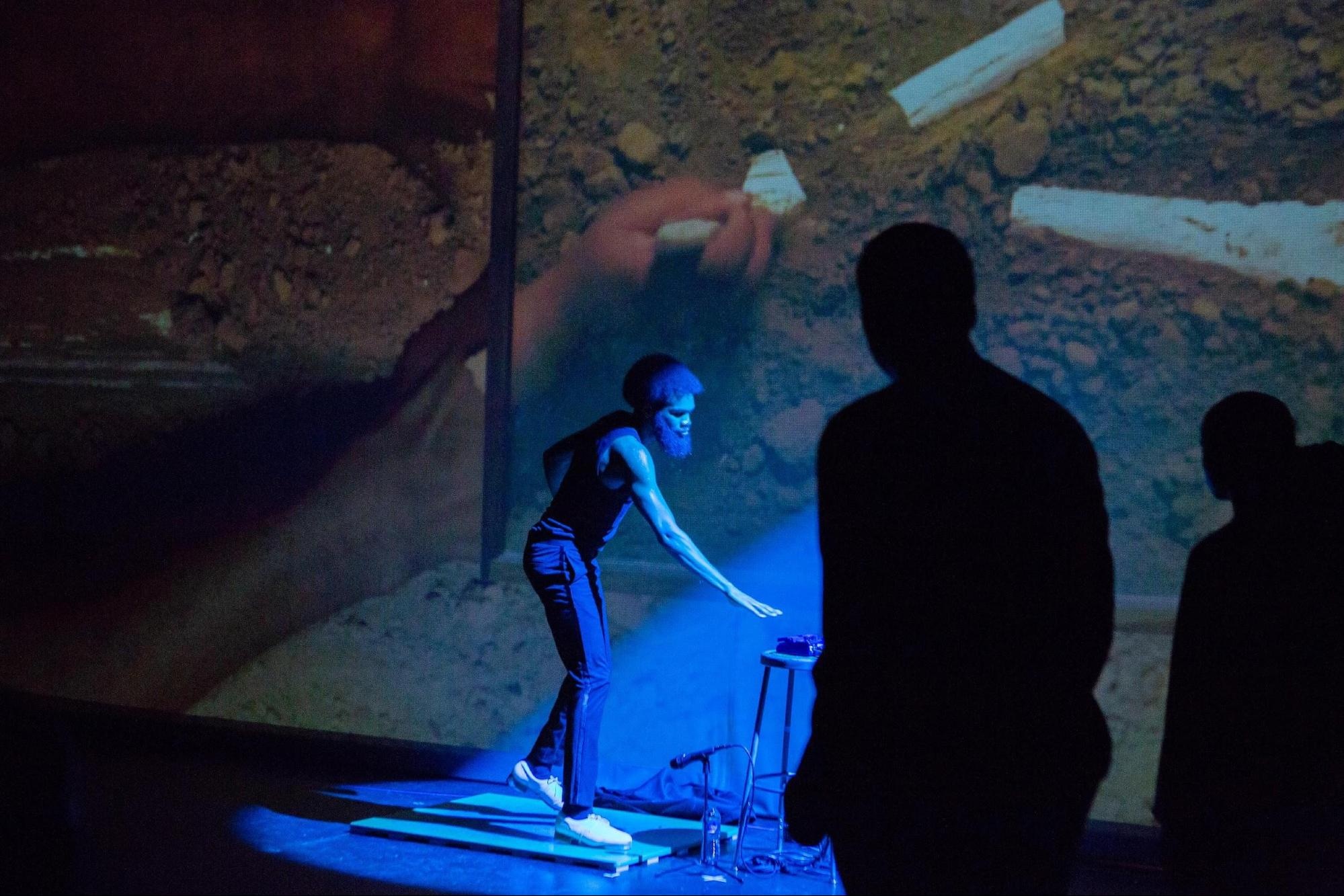 Dancer Michael J. Love, a Black man wearing a tank top, slacks, and white sneakers, stands in a pool of blue light. 