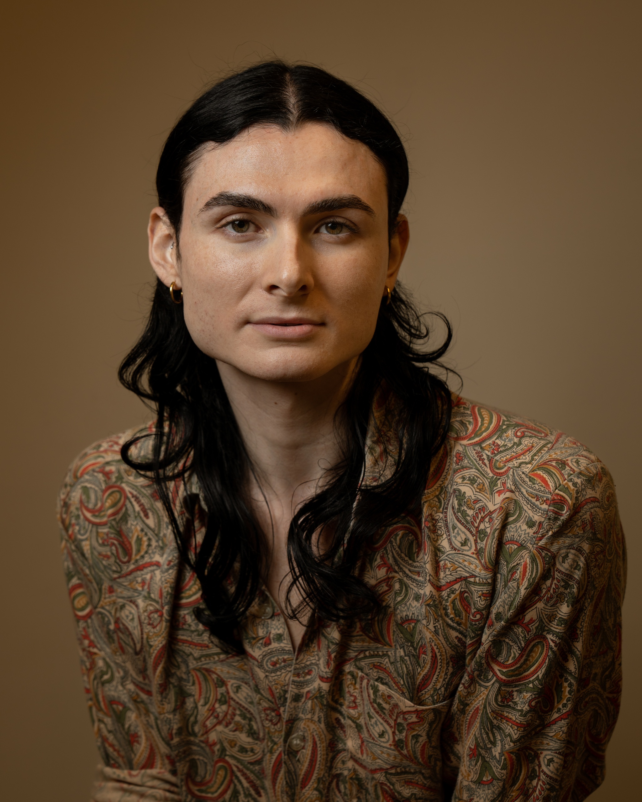 A portrait of Joshua Leon Eguia who poses against a neutral brown background. Joshue has long dark brown wavy hair that is tied back and falls to shoulder length. Joshua wears an earth-toned paisley print shirt and looks directly at us.