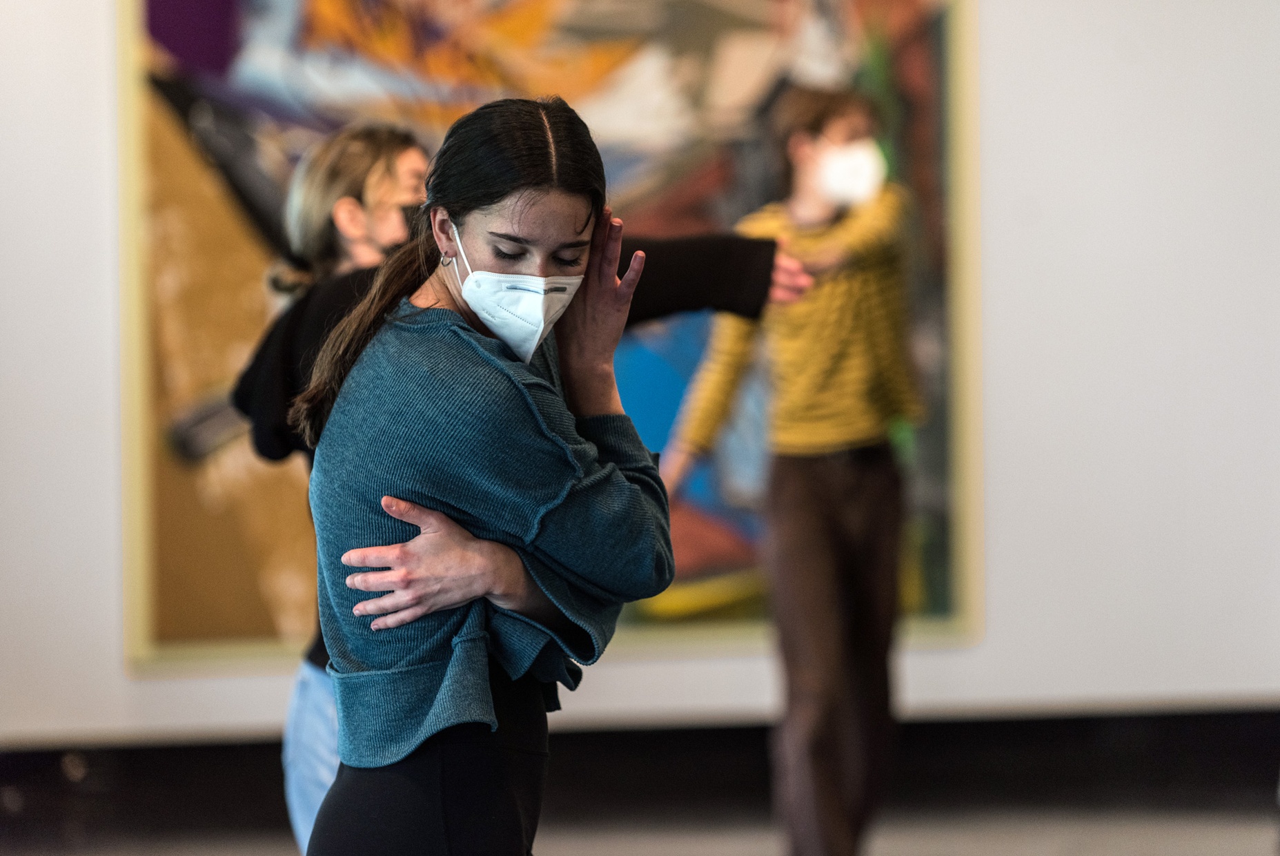 A masked young woman poses while dancing in front of an abstract painting and two other people who are out of focus behind her.