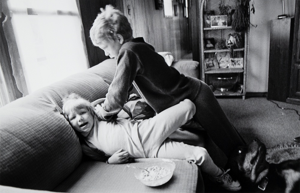 A black and white photograph of a boy looking down onto and pressing a girl into a couch as she looks at the camera.