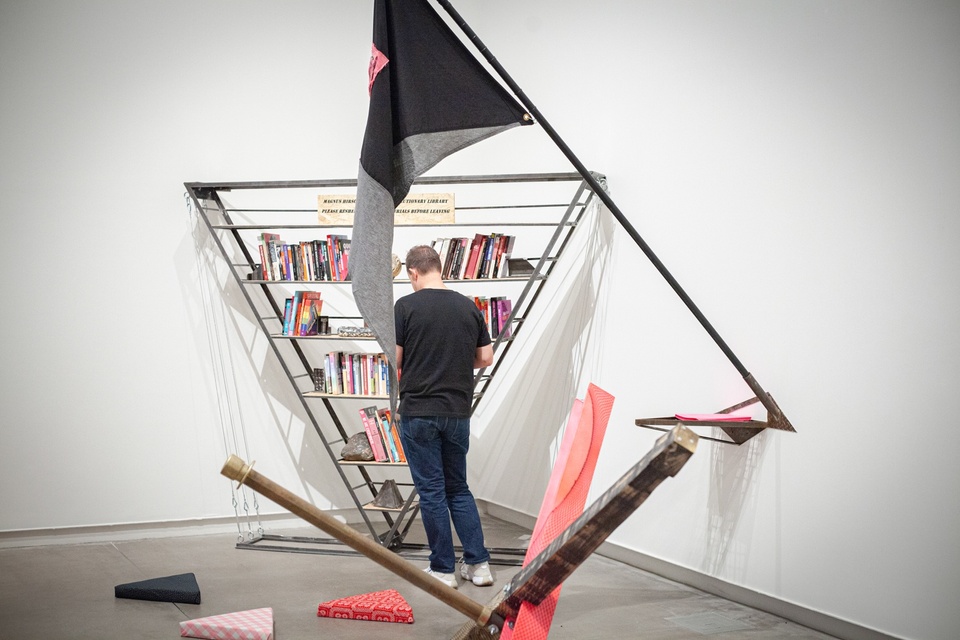 Person reads a book from a metal bookshelf constructed in an upside-down triangle shape. A flag made of black, gray, and pink fabric scraps is mounted on the wall behind them.