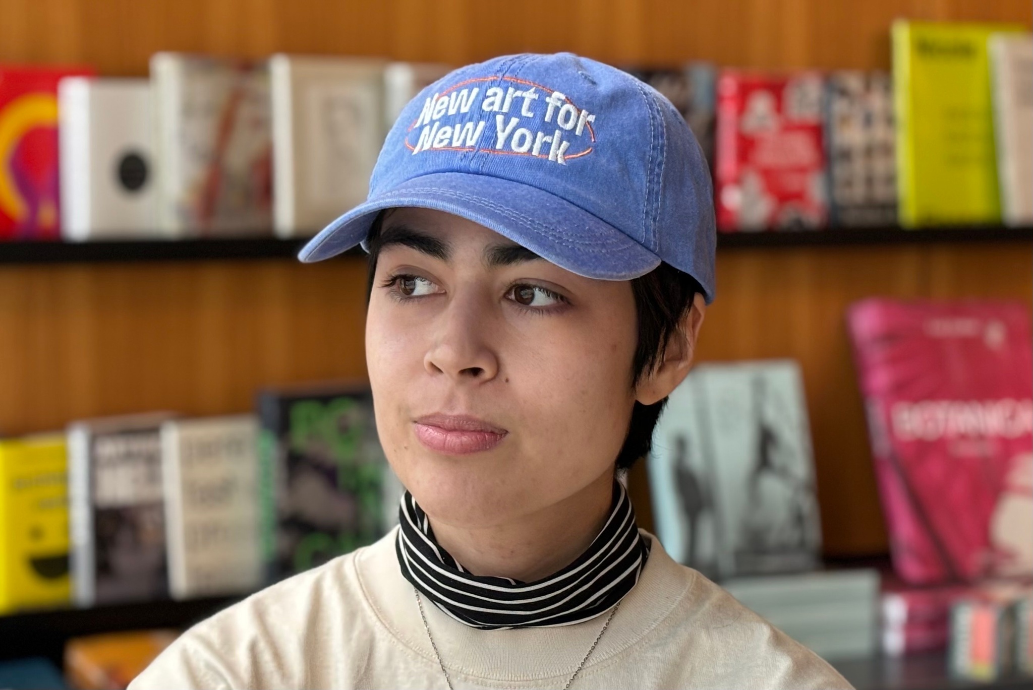 A model wears an Open Call baseball cap. The cap is a light blue, like a periwinkle denim. Across the front of the cap is embroidered in white letters: New art for New York. Behind the model are rows of books softly out of focus and facing outward along a wooden wall.