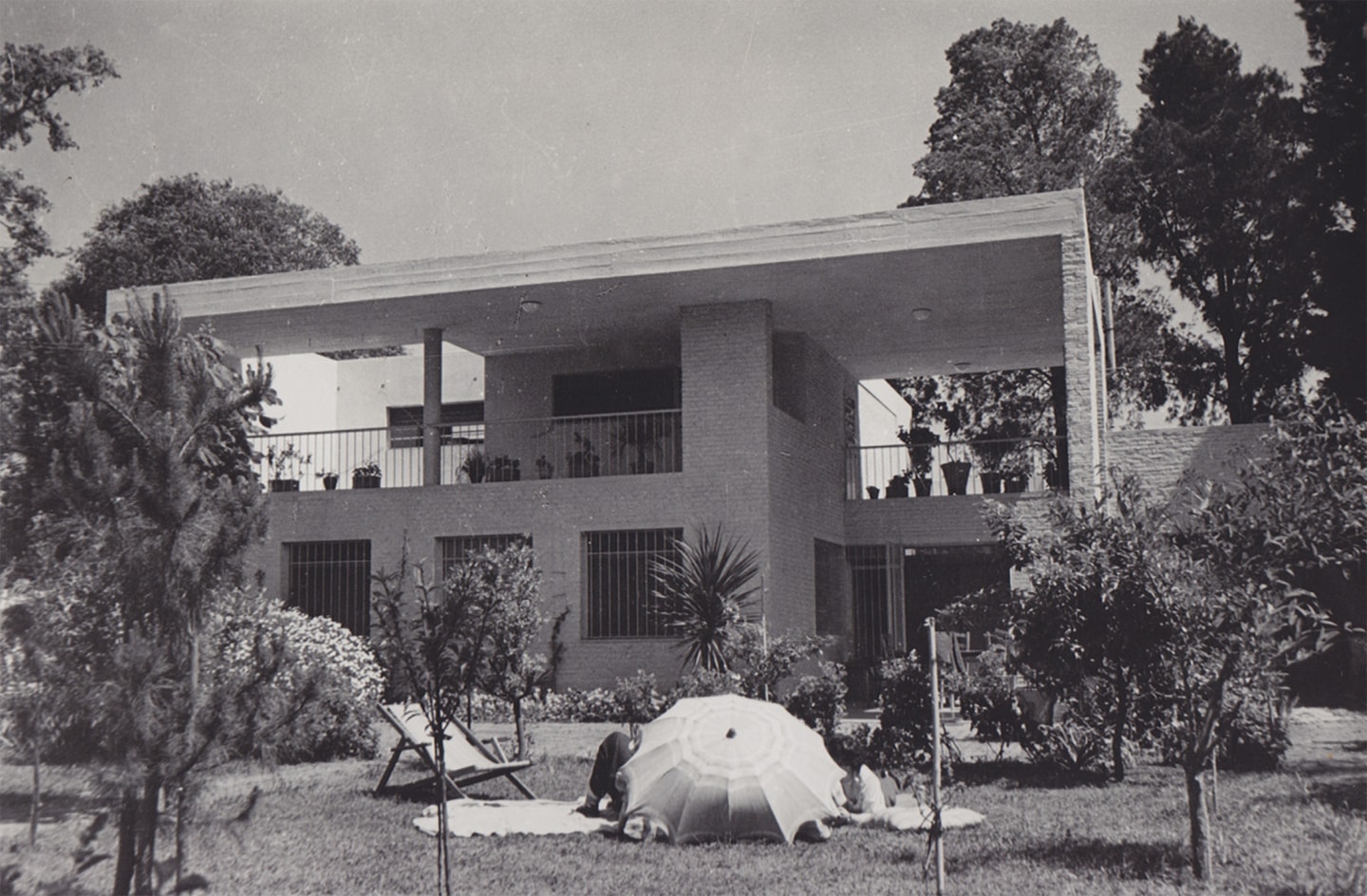 Black-and-white photograph of a two-story modernist house designed by Wladimiro Acosta for Horacio Coppola and Grete Stern, with a yard and trees in the foreground and people reclining on the lawn beneath an umbrella.