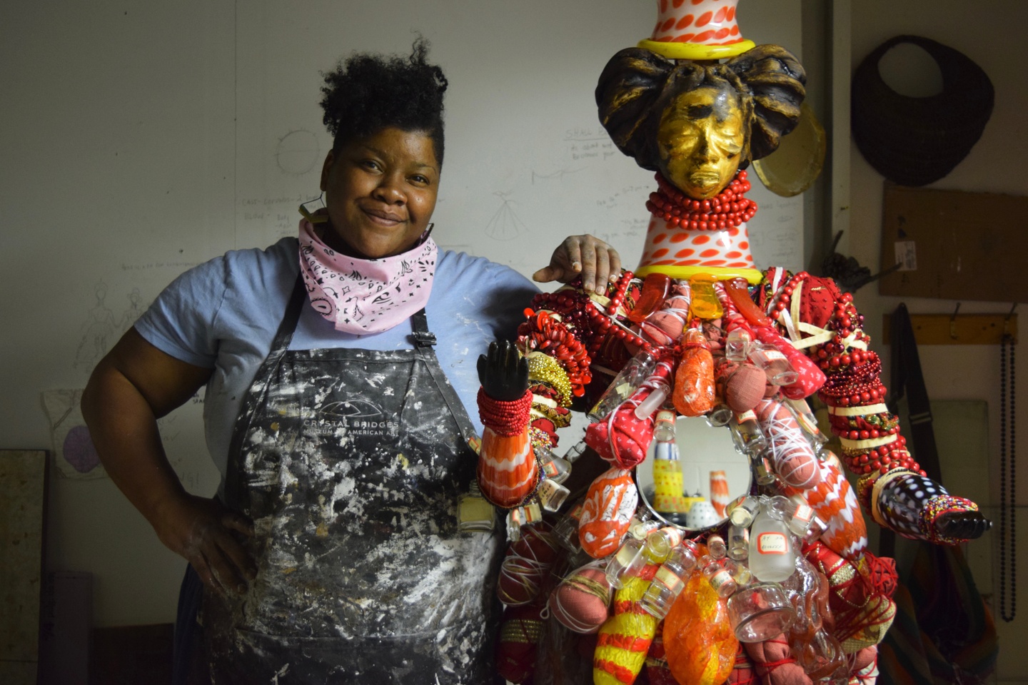 Vanessa German standing next to a sculptural figure of a person she has created with an assortment of materials—beads, jars, a small round mirror. The body of the figure is primarily red; the head is golden, with two large buns and a hat.