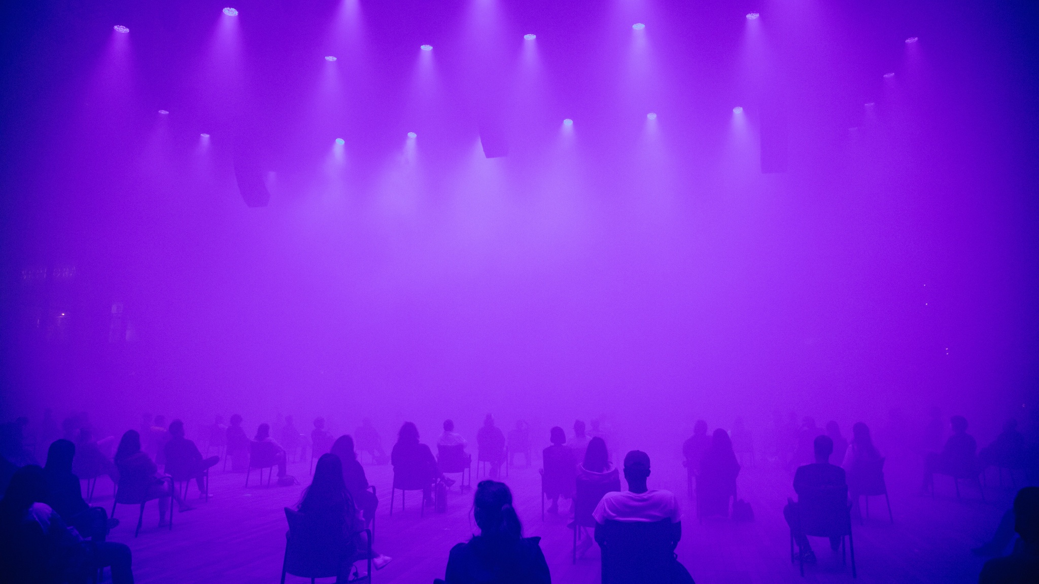 An audience seated in distanced seats in a foggy performance space lit with diffuse purple lighting