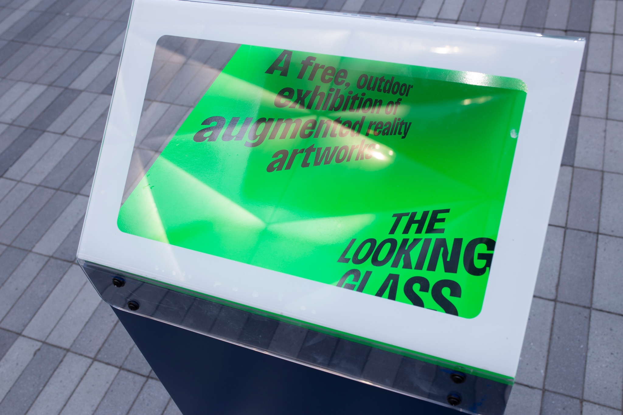 A green label with the words "The Looking Glass, A free outdoor exhibition of augmented reality artworks" under a transparent plastic cover that reflects some light