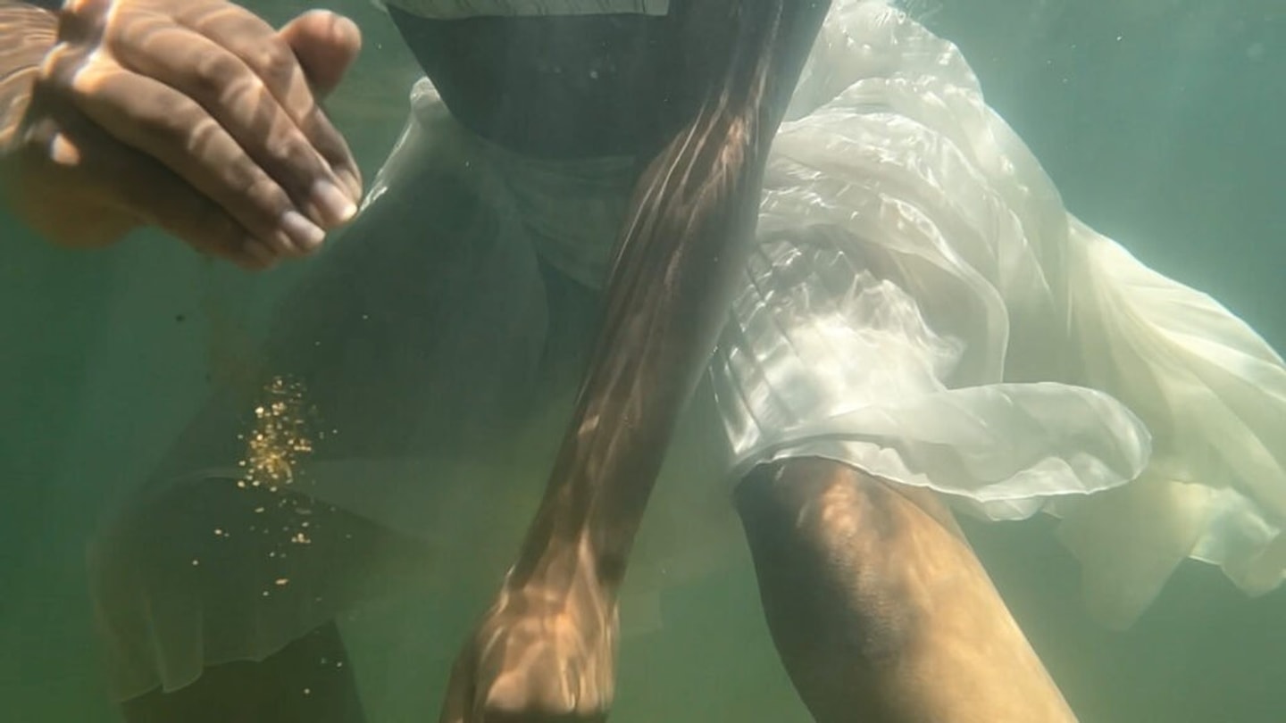 still from a video, close up of arms and legs in a white dress underwater