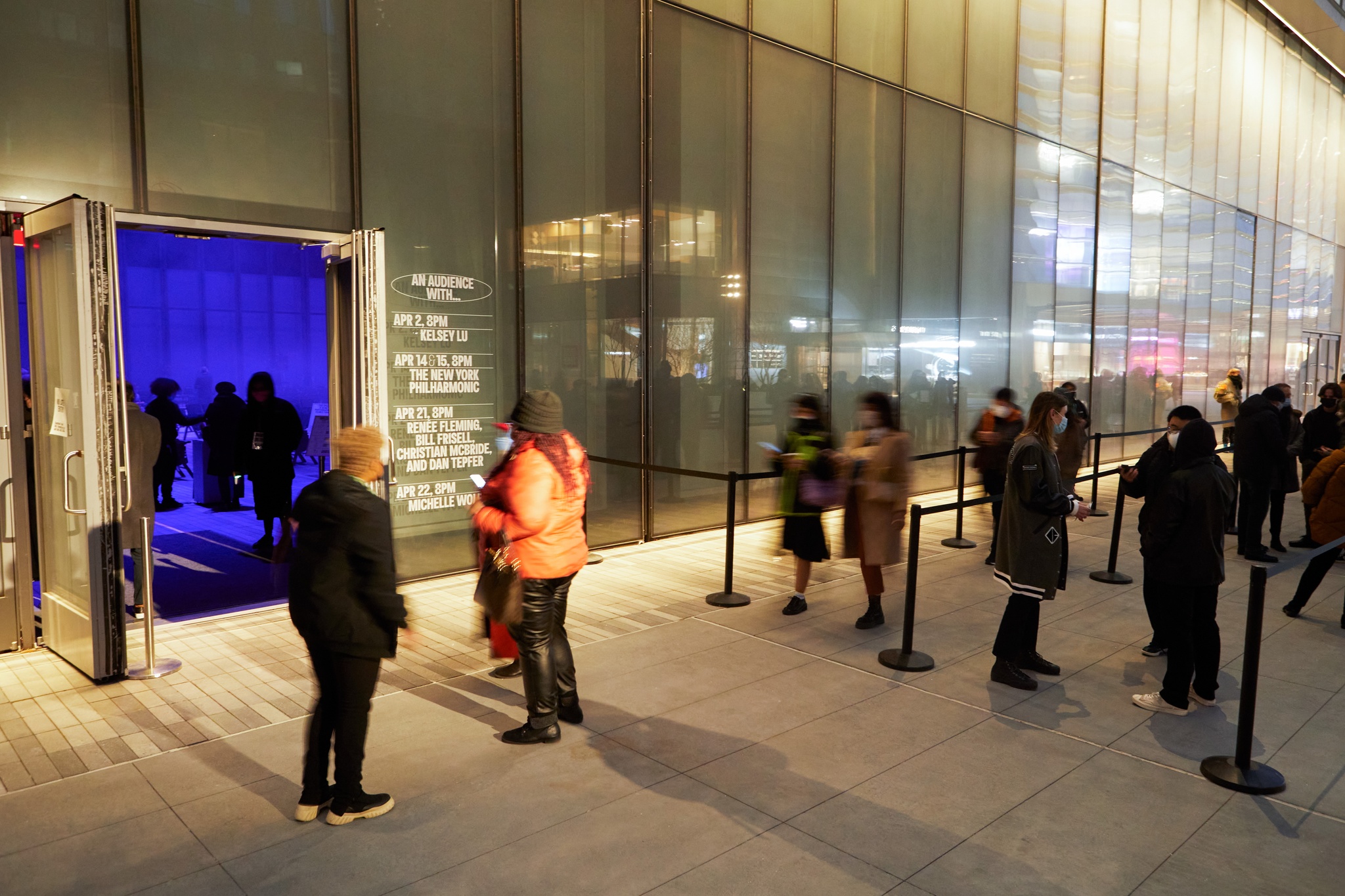 People in a stanchioned line outside The Shed at night. An open door reveals a blue-lit performance space inside.