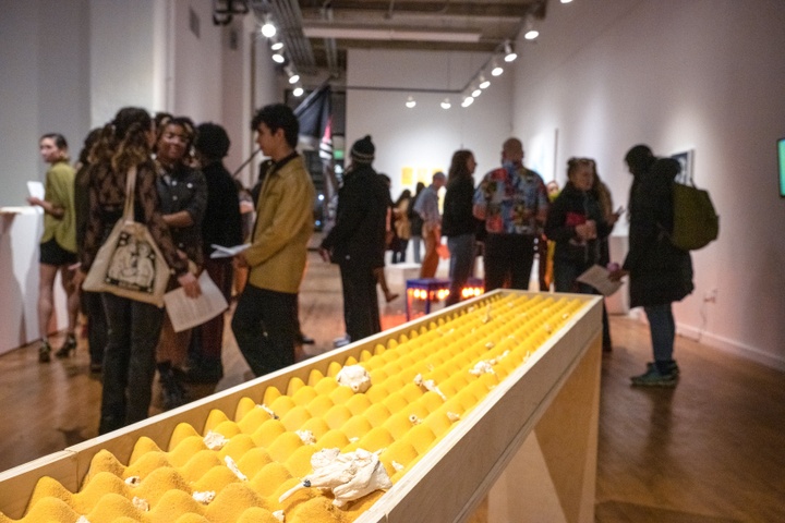 Gallery space full of visitors. In the foreground is a sculpture set into a table consistent of a bright yellow egg-crate mattress pad littered with small plaster objects.