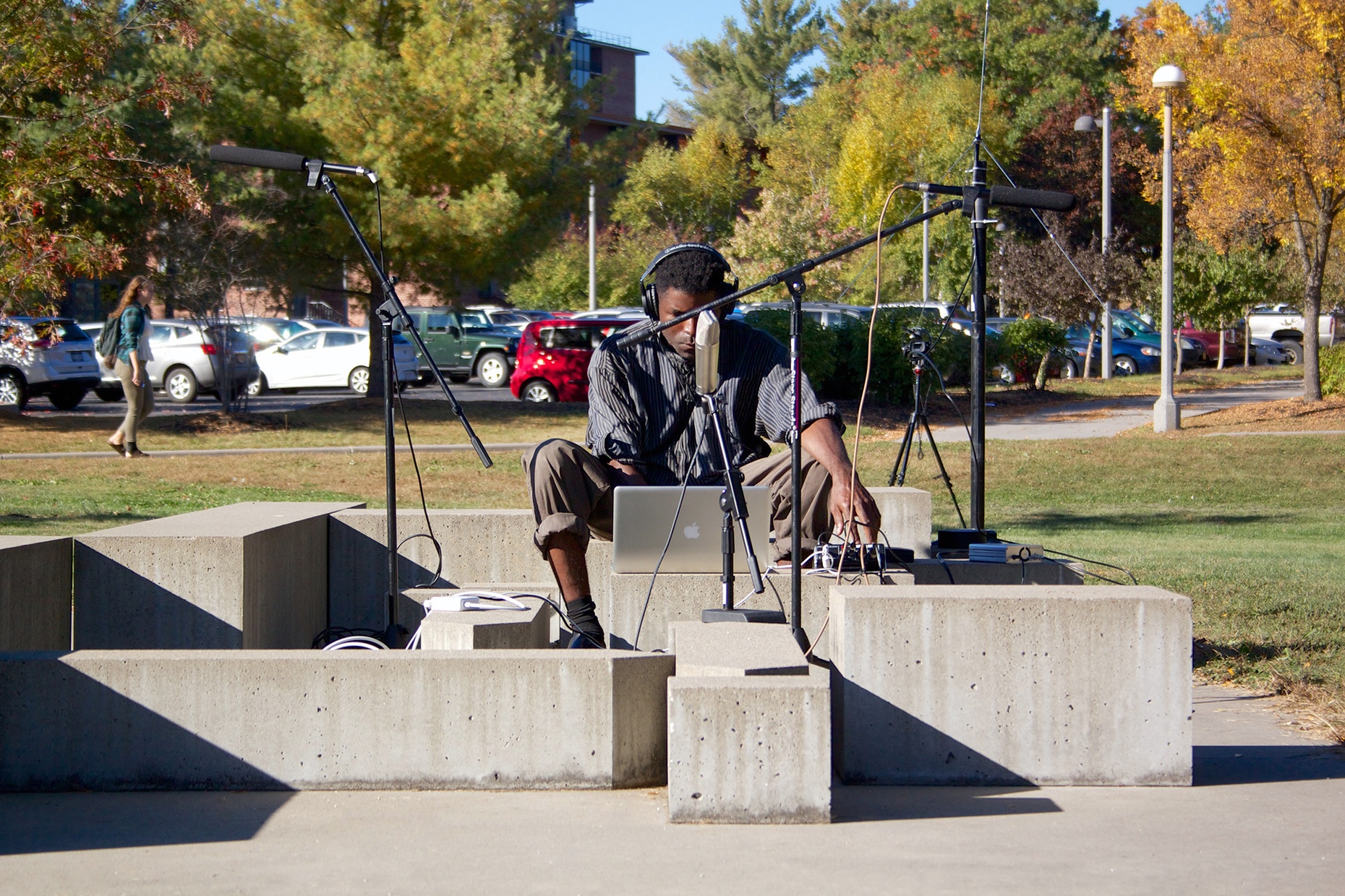 A black man sits on an outdoor sculpture made of concrete blocks with a laptop and two mics on stands.