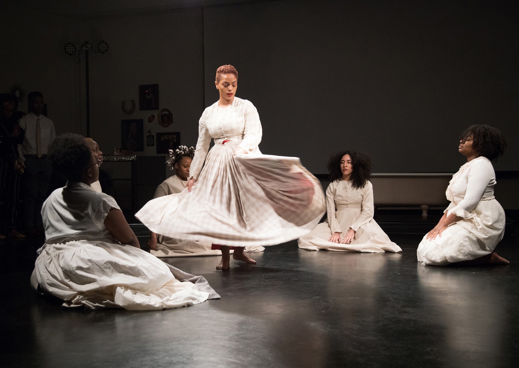 Six women of color are performing in a dark room with a black floor. Five of the women sit on the ground in a circle while one woman stands towards the center and twirls her skirt. They are all wearing white dresses. 