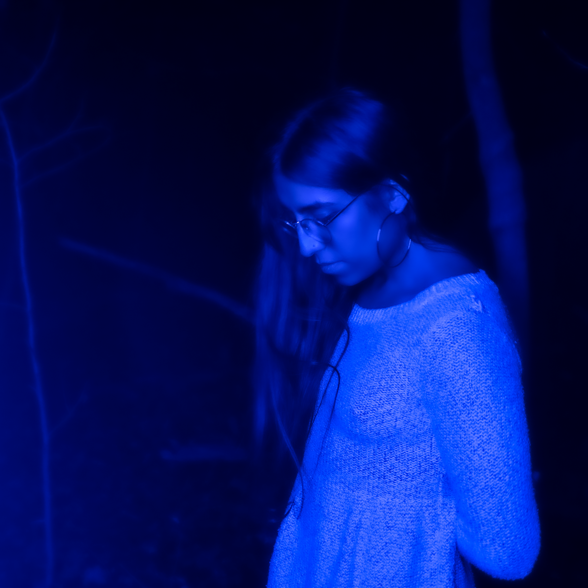 A photo of artist Rachika Nayar bathed in blue light. Rackika is looking down with her long hair falling straight down the right side of her head and torso.