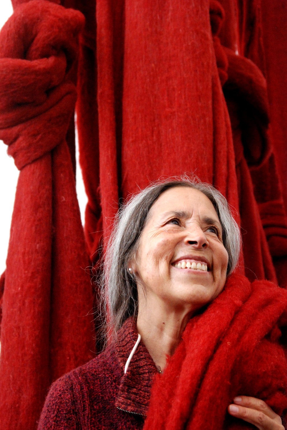 Poet, activist, and artist Cecilia Vicuna looks upward with a thick skein of bright red wool draped over her shoulder. She smiles and has long gray hair pulled back loosely behind her head. Behind her broad swaths of the red fabric hang from above. One of these swaths has a large knot tied in it.