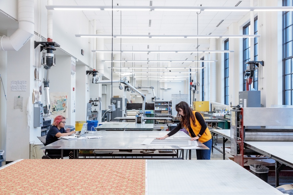 Spacious, light-filled printmaking studio space; two students are working at a table in the middle of the room.