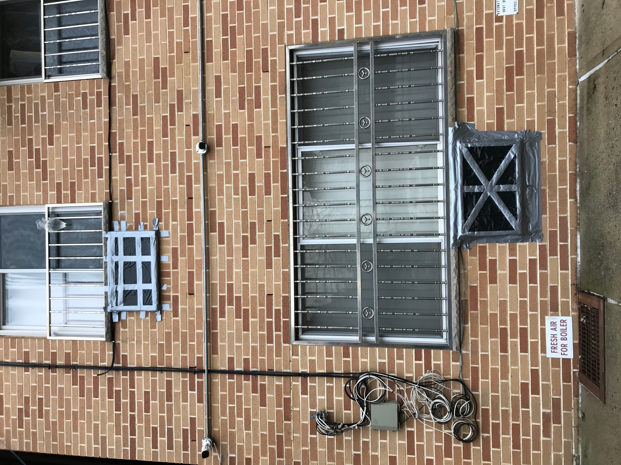 The side of a brick apartment building. The first story has a wide, three-paned window with stainless steel bars over it. Areas cut out for A/C units on the first and second stories are covered in black plastic tarp sealed with crisscrossing duct tape.