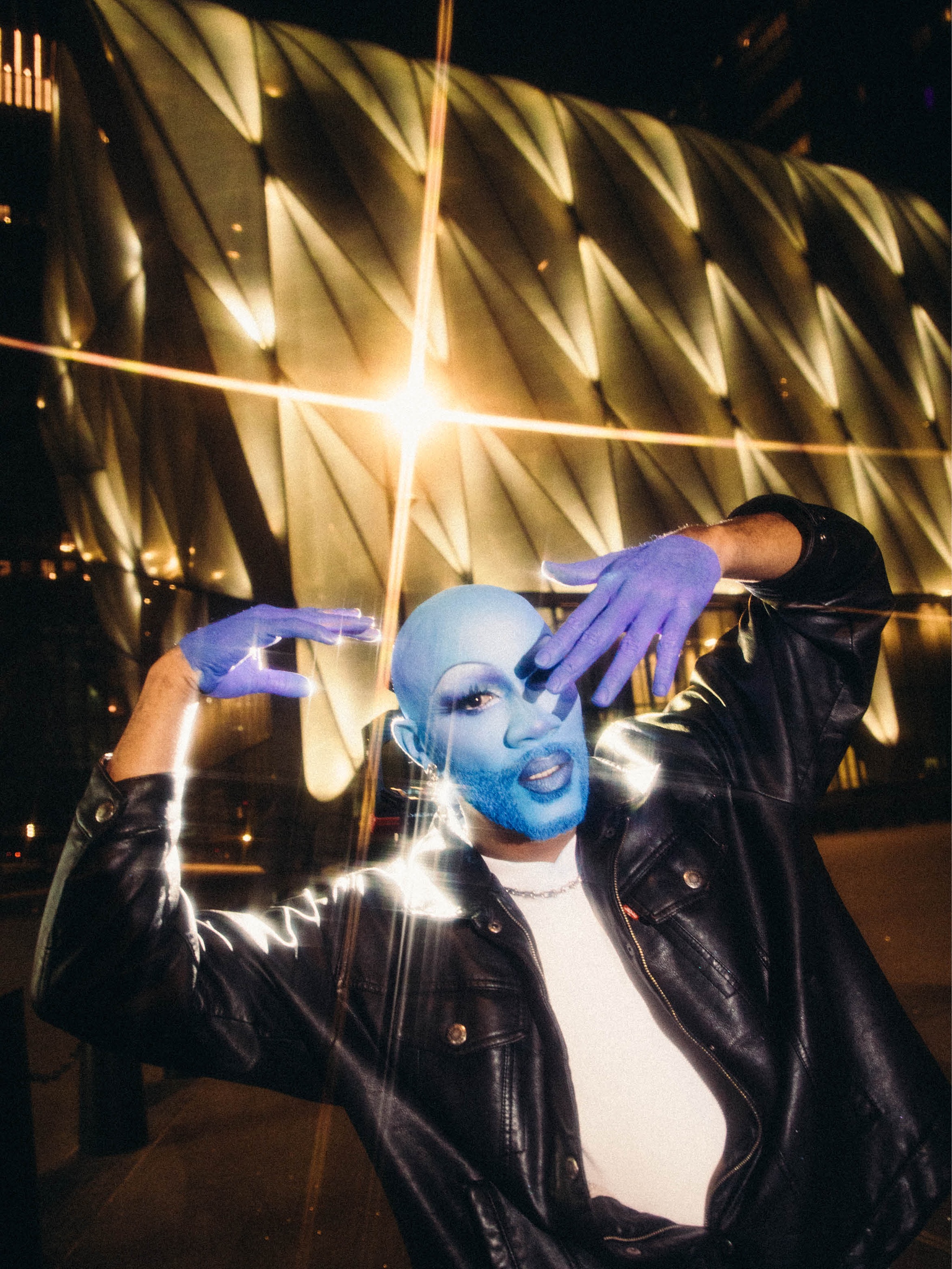 Cain Coleman, a Black multihyphenate drag performer, poses at night outside The Shed with their head and face covered in blue makeup. Cain wears a black leather jacket and white t-shirt, holding their hands, covered in purple makeup to resemble gloves, up to either side of their face. Behind them, lights flash off the reflective surface of The Shed in small starbursts. 