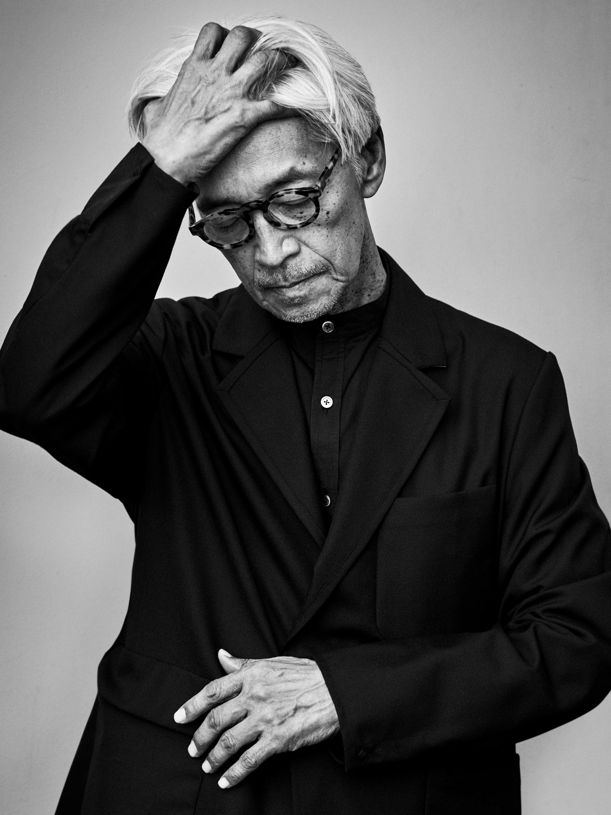 A black and white portrait of an older Japanese man with gray hair. He raises one hand to push his hair back and casts his gaze downward, holding his other hand flat against his waist. He wears dark clothes and glasses. 